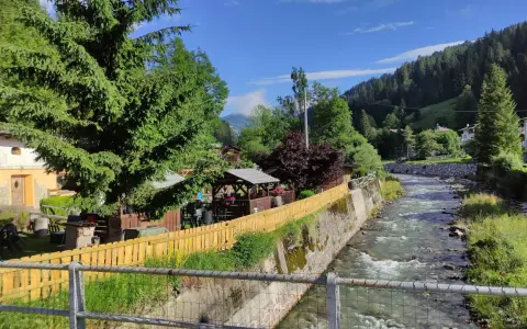 Un ruscello scorre a Santo Stefano di Cadore, fiancheggiato da case, alberi e un'area con gazebo e tavoli all'aperto.