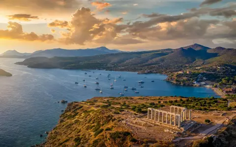 Rovine del Tempio di Poseidone a Capo Sounion, con barche a vela che navigano nel mare blu sotto un cielo dorato al tramonto.