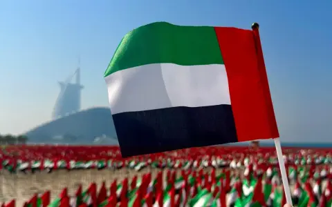 Flaga Zjednoczonych Emiratów Arabskich powiewa na tle drapaczy chmur w Dubaju. W tle widać morze małych flag.