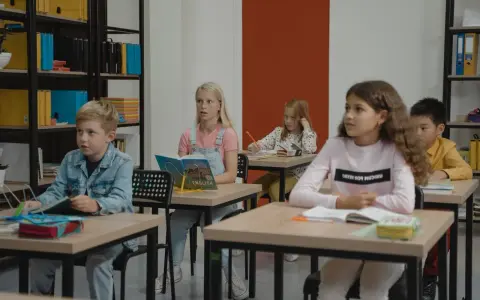 Dzieci siedzą w klasie, skupione na lekcji. Chłopiec w dżinsowej kurtce, dziewczynka z książką po angielsku, a z tyłu inne dzieci. Wszyscy mieli nadzieję, że lekcja szybko się skończy.