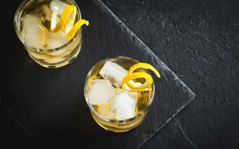 Zwei erfrischende Whiskey Cocktails mit Eis und Zitronenzesten auf dunklem Stein.