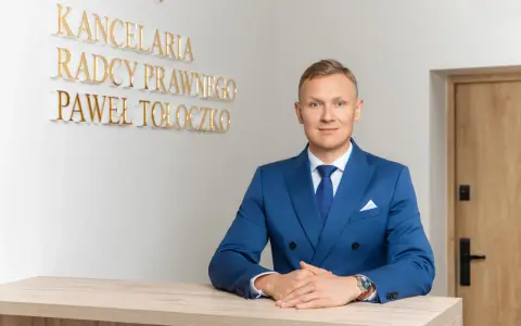 Mężczyzna w niebieskim garniturze przy biurku, za nim napis "KANCELaria Radcy Prawnego Paweł Tołoczko".