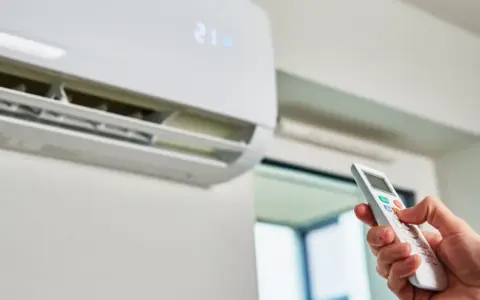 Mano con control remoto ajustando el aire acondicionado a 21 grados. ¿Es mejor apagar el aire acondicionado o dejarlo encendido?