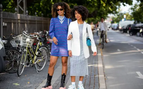 Two women showcase their spring chic dress code. One wears a blue plaid dress and chunky boots, the other a white blazer over a patterned dress and sneakers.