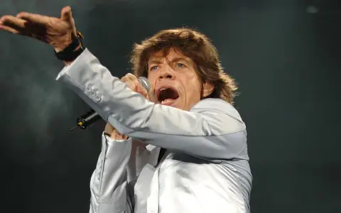 Mick Jagger, wokalista zespołu, śpiewa z mikrofonem, gestykulując ręką.