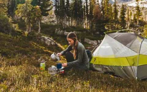 A woman enjoys a peaceful moment of camping alone, preparing a warm drink next to her tent amidst a serene forest landscape.