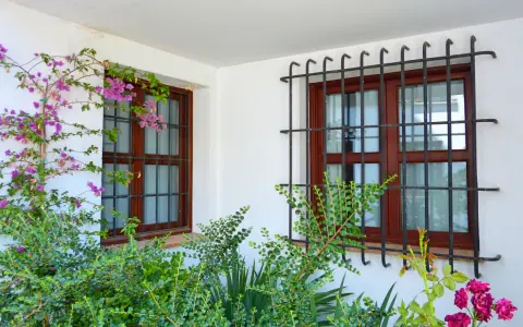 Dos ventanas con barrotillo de hierro y marcos de madera, rodeadas de vegetación y flores moradas.