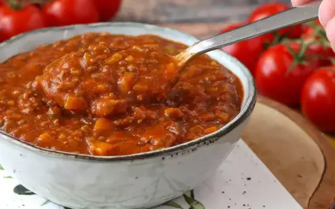 Une cuillère prélève une portion généreuse de sauce bolognaise maison, riche et savoureuse, dans un bol. Des tomates fraîches en arrière-plan.