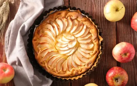 Tarta de manzana dorada, decorada con láminas de fruta, rodeada de manzanas frescas y espigas de trigo sobre una mesa de madera.