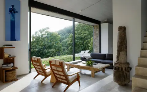 Salón moderno con sofás, sillones y mesa de centro rústica. Los suelos de piedra, con un estilo que evoca los años 70, contrastan con la vista exterior de un bosque frondoso.