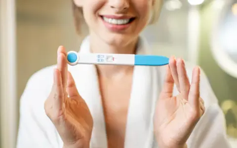 Uśmiechnięta kobieta w szlafroku trzyma test ciążowy z dwoma kreskami. To moment, kiedy warto zastanowić się, kiedy zrobić test ciążowy.