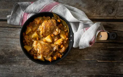 Estofado de pollo con patatas y zanahorias en sartén sobre mesa de madera.