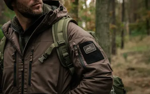 Mężczyzna w brązowej kurtce z naszywką Helikon-Tex, symbol polskie marki sportowe, w lesie.