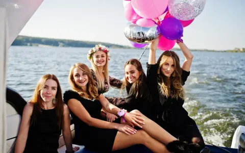 Uśmiechnięte kobiety na łodzi z balonami, celebrujące organizację wieczoru panieńskiego.