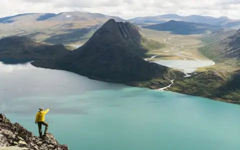 En person i gul jacka pekar ut över en turkos sjö och berglandskap under en Jotunheimen vandring.