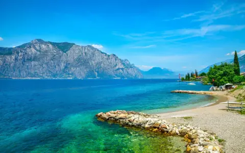 Ein idyllischer Gardasee Strand mit kristallklarem Wasser, felsigen Ufern und majestätischen Bergen im Hintergrund.