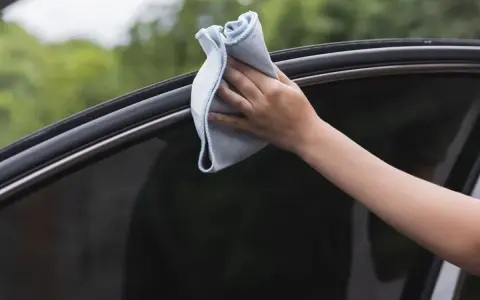 Mano limpiando los cristales del coche con un paño de microfibra azul. Un paso esencial para saber cómo limpiar los cristales del coche y mantener una visibilidad perfecta.