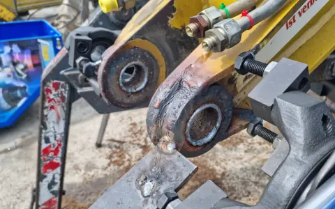 Naprawa tulejowania koparki. Widoczne elementy hydrauliczne i zardzewiałe sworznie.