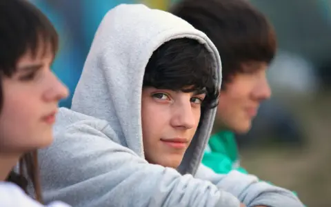 Drei Jungs, etwa 13 Jahre alt, sitzen nebeneinander. Einer trägt einen grauen Kapuzenpullover, der sein Gesicht fast verdeckt.