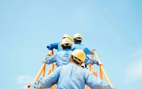 Trabajadores con cascos amarillos suben una escalera naranja contra un cielo azul claro. Podría ser una escena de cesión ilegal de trabajadores.
