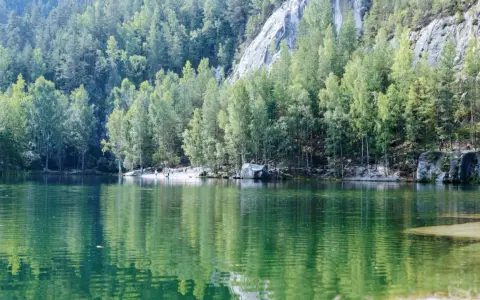 Zielona tafla jeziora odbija las i skalne miasto w Polsce. Ludzie spacerują po piaszczystym brzegu.