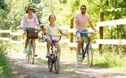 Rodzina cieszy się rodzinnym wypadem na **Mazury rowerem**. Radosna dziewczynka jedzie na fioletowym rowerze, a za nią rodzice.