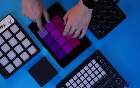 Des mains manipulent une tablette affichant des pads colorés, entourée de contrôleurs midi. Création musicale en cours.