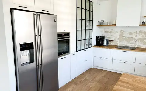 Nowoczesna kuchnia z białymi frontami szafek w odcieniu **ral 9016**, lodówką side-by-side i czarną, industrialną witryną.