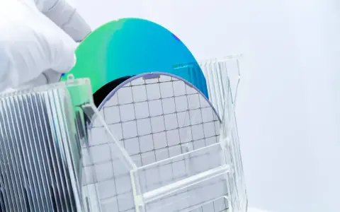 A gloved hand holds a silicon wafer, its grid pattern visible. Another wafer, iridescent blue and green, rests behind it. This image explores the question: is plastic a semiconductor?