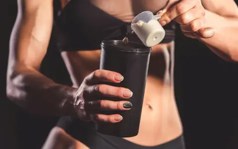 Kobieta z umięśnionym brzuchem dodaje proszek do shakera. Czy shake proteinowy jest zdrowy? To zależy od składu.