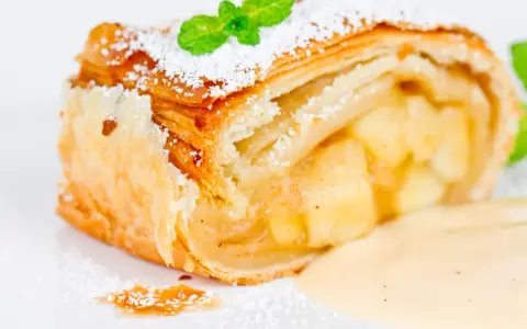 Ein Stück Apfelstrudel mit Filoteig, bestäubt mit Puderzucker und garniert mit Minze, serviert mit Vanillesauce.