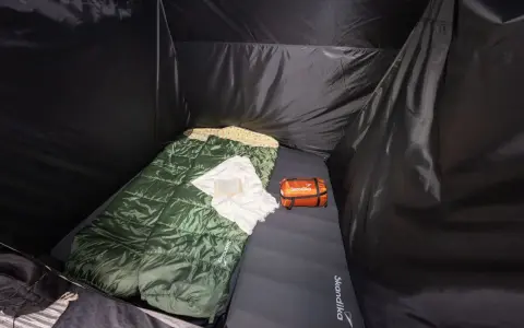 Schlafsack und Kissen auf der Isomatte, welche Seite oben ist, ist klar erkennbar. Ein kleines orangefarbenes Päckchen liegt daneben.