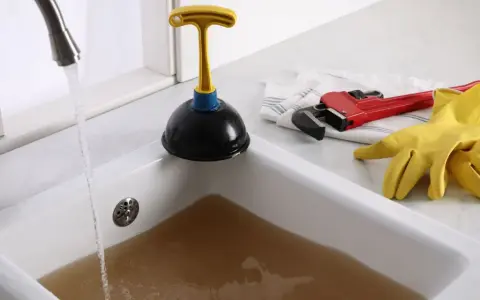 Un évier bouché après siphon, avec un ventouse, une clé à molette rouge et des gants jaunes prêts à l'emploi. L'eau sale s'accumule.