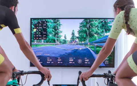 Two cyclists race virtually on a scenic road using indoor cycling apps. The screen displays their stats and rankings.
