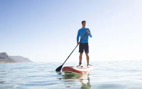 Mann steht auf SUP-Board, hält Paddel richtig. Ruhige See, bergige Küste im Hintergrund.