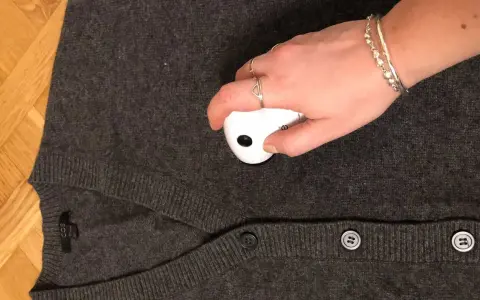 En hand med smycken använder en noppmaskin på en mörkgrå kofta. Materialet känns mjukt, kanske rayon material svenska.