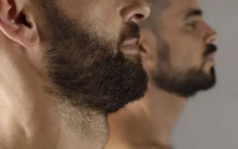Primer plano de un hombre con barba oscura y tupida, reflejado en un espejo. La imagen captura la decisión de dejar crecer la barba.