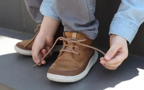 Kind lernt, seine braunen Schnürstiefel zu binden. Die Schnürsenkel sind hellbraun und gemustert.