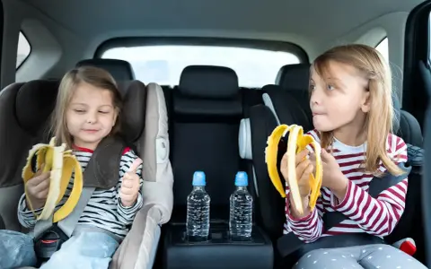 Dwie dziewczynki w fotelikach samochodowych jedzą banany. Woda i przekąski to podstawa, gdy zastanawiasz się, co zabrać na długą podróż samochodem z dzieckiem.