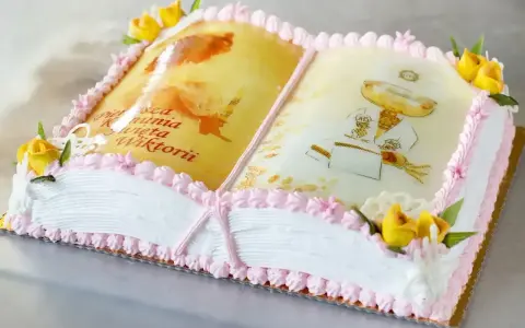 Eleganckie ciasto na komunię w kształcie otwartej księgi z motywami religijnymi i napisem "Pierwsza Komunia Święta Wiktorii".