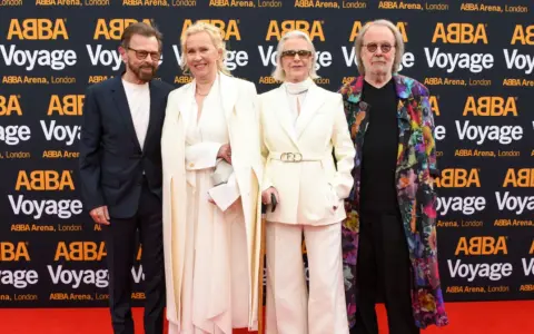 Członkowie zespołu ABBA na czerwonym dywanie, pozujący do zdjęć przed banerem "ABBA Voyage".