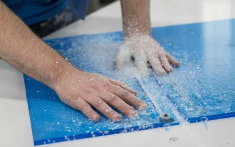 Hands guide a router, creating a clean cut on a blue plexiglass sheet, leaving a trail of white shavings. This is how you cut plexiglass with a router.