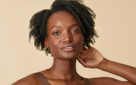 Une femme à la peau foncée, aux cheveux afro volumineux et bouclés, porte des boucles d'oreilles dorées.