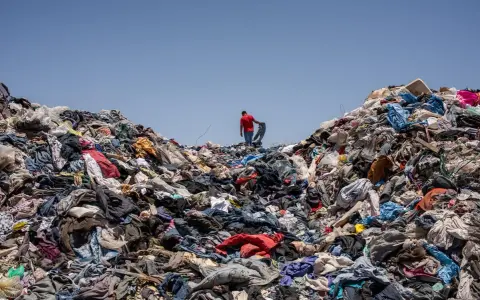 Un uomo in maglia rossa cammina su una montagna di vestiti scartati, simbolo dell'inquinamento tessile.