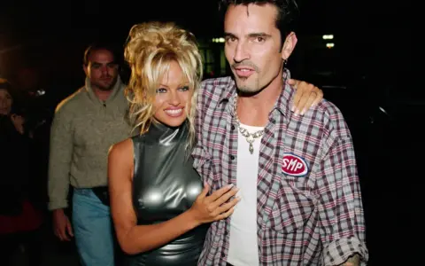 Pamela Anderson i Tommy Lee, ikony lat 90., pozują do zdjęć. Pamela w błyszczącej sukience, Tommy w kratowanej koszuli.