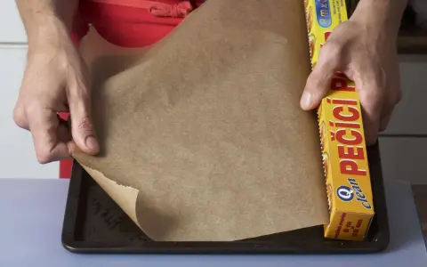 Hände legen Backpapier auf ein Blech. Die Rolle mit der Aufschrift "PEČÍCÍ PAPÍR" zeigt, dass man Backpapier einfetten muss, um ein Anhaften zu verhindern.