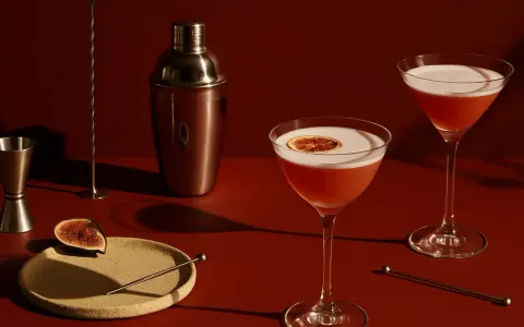 Elegancki drink z czerwonym winem, ozdobiony plasterkiem cytrusa, obok shakera i kieliszka z pianką.
