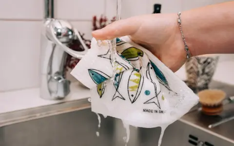 En hand sköljer en disktrasa med fiskmönster under rinnande vatten. Trasan är gjord av garn till disktrasa och har texten "Made in Sweden".