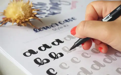 Mano practicando cómo hacer lettering con un rotulador sobre un papel con letras de ejemplo.