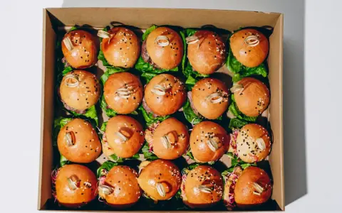 Pudełko z dwudziestoma mini burgerami na liściach sałaty, posypane sezamem i ozdobione patyczkami.